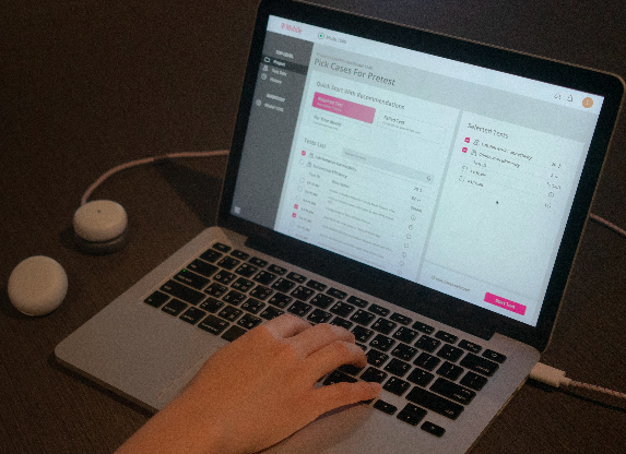 A person is using a laptop featuring a software interface titled Pick Cases for Pretest. The screen displays options and checkboxes. Two small, white, oval devices are on the desk beside the laptop, with a hand interacting with the keyboard.