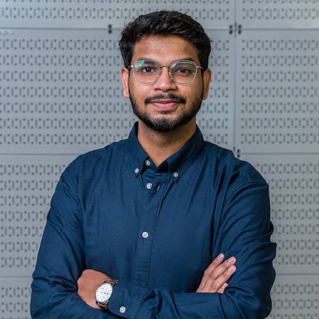 A man with short hair and a beard is wearing glasses and a dark button-up shirt. He stands with his arms crossed, smiling slightly against a backdrop of a light gray patterned wall.