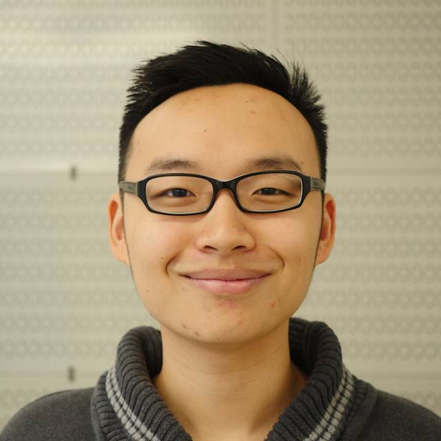 A person with short black hair and black-rimmed glasses smiles at the camera. They are wearing a gray sweater with a patterned collar. The background is a light-colored wall with a subtle geometric pattern.
