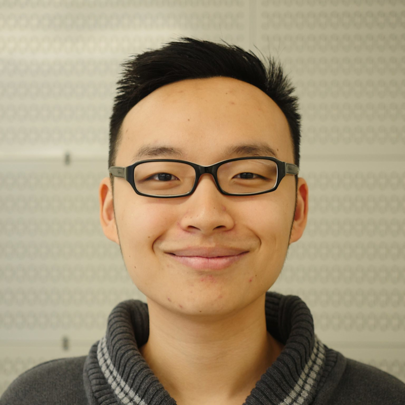 A person with short black hair and black-rimmed glasses smiles at the camera. They are wearing a gray sweater with a patterned collar. The background is a light-colored wall with a subtle geometric pattern.