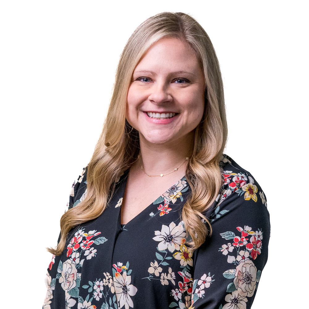 A person with long blonde hair smiles while wearing a black cardigan over a red blouse with floral patterns. The background is a light gray wall with a pattern of small squares.