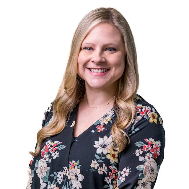 A person with long blonde hair smiles while wearing a black cardigan over a red blouse with floral patterns. The background is a light gray wall with a pattern of small squares.