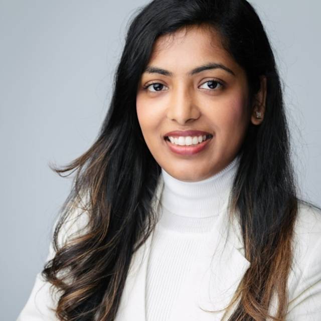 Smiling person with long dark hair wearing a white turtleneck and blazer against a plain background.