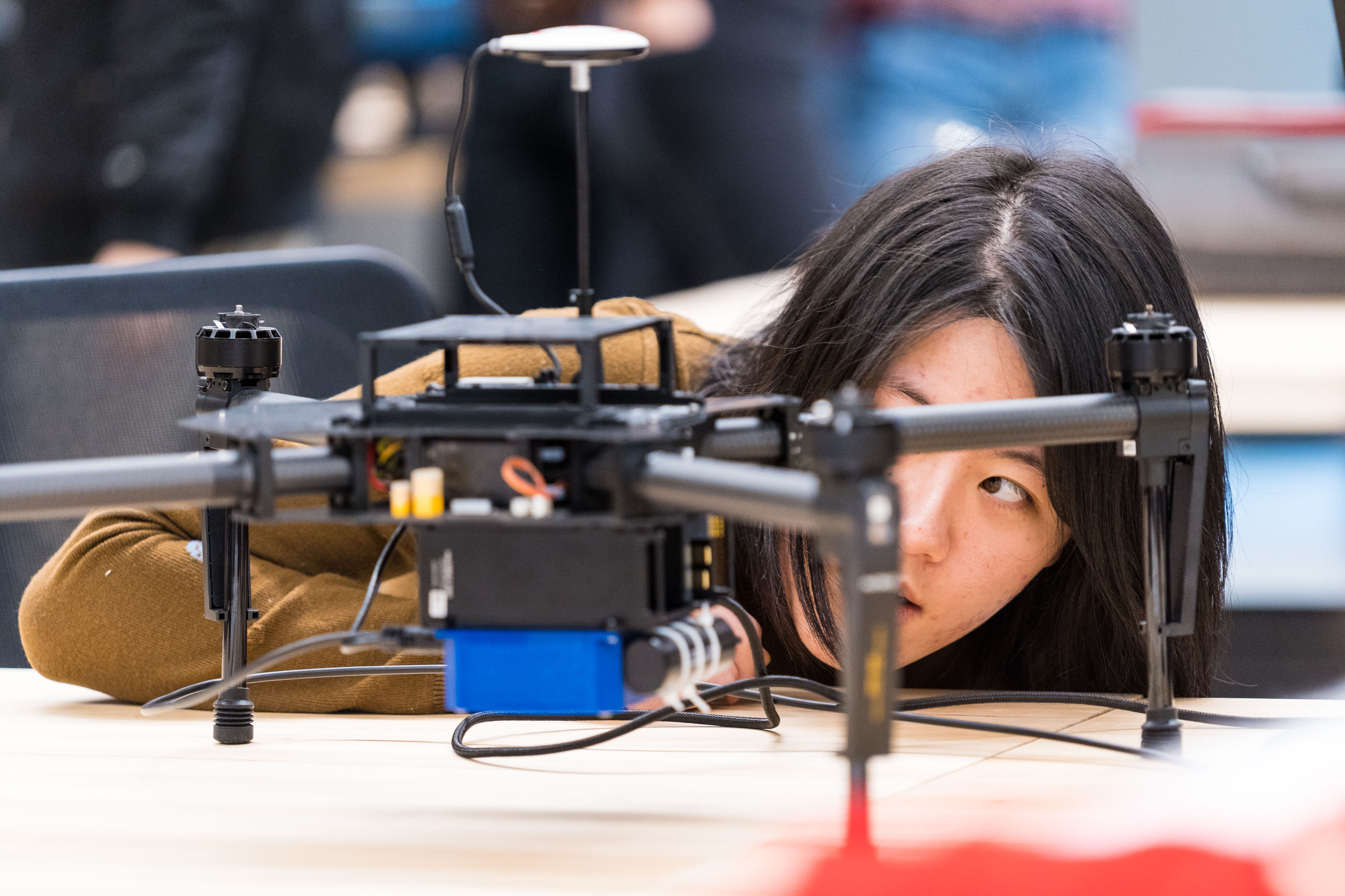 A woman closely examines a drone placed on a table. She is focused on its details, with her face partially visible through the drones structure. The background is blurred, suggesting an indoor setting with other people present.