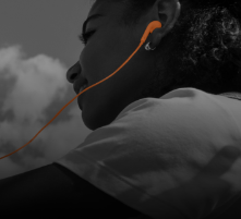 A person wearing orange earphones, looking upward. The background shows a cloudy sky. The image is mostly in grayscale except for the orange earphones.