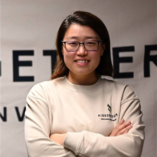 Person with shoulder-length hair and glasses, wearing a beige sweatshirt with RISE2GETHER logo, stands smiling with crossed arms. The background shows a partially visible text banner.