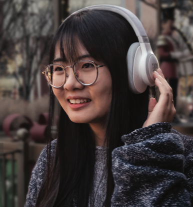A person with long dark hair and glasses is smiling while wearing white over-ear headphones, standing outdoors with a blurred background of trees and buildings.