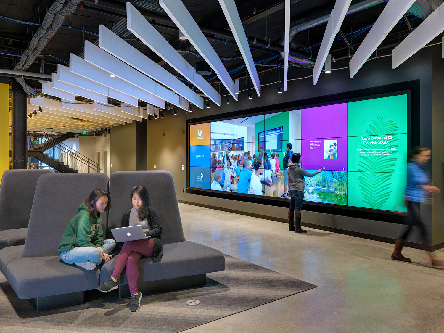 Two people sit on modern gray seating using a laptop, while a third person interacts with a large, colorful digital display on the wall in a sleek, industrial-style room with overhead lighting.