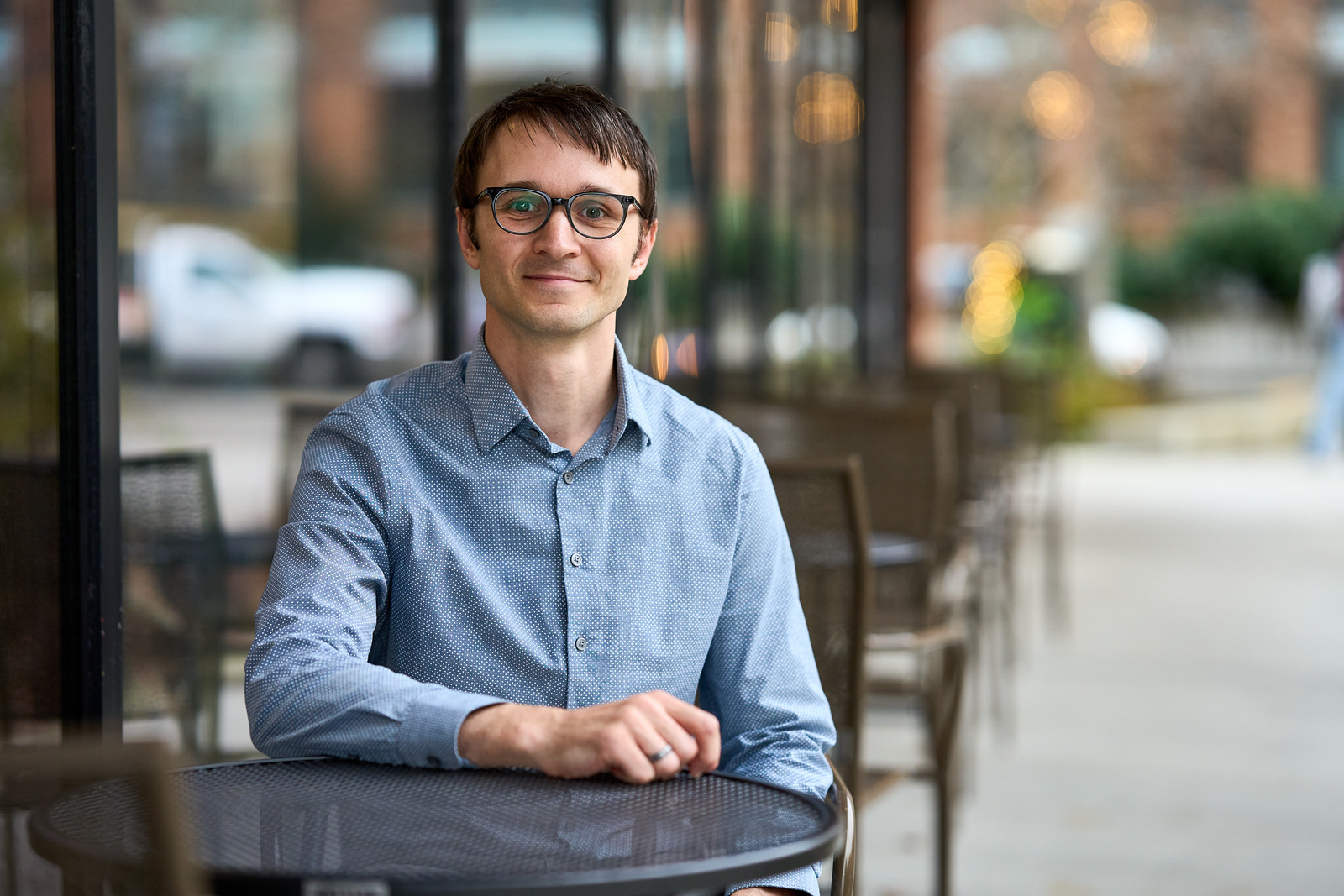 A man with glasses and a blue shirt sits at an outdoor café table. He is smiling and looking at the camera. The background shows a blurred street scene with buildings and lights.