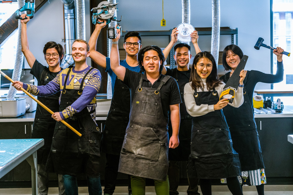 A group of seven people stand in a workshop, smiling and holding various tools. They are wearing black aprons. The background shows workbenches and equipment, suggesting a collaborative or creative environment.