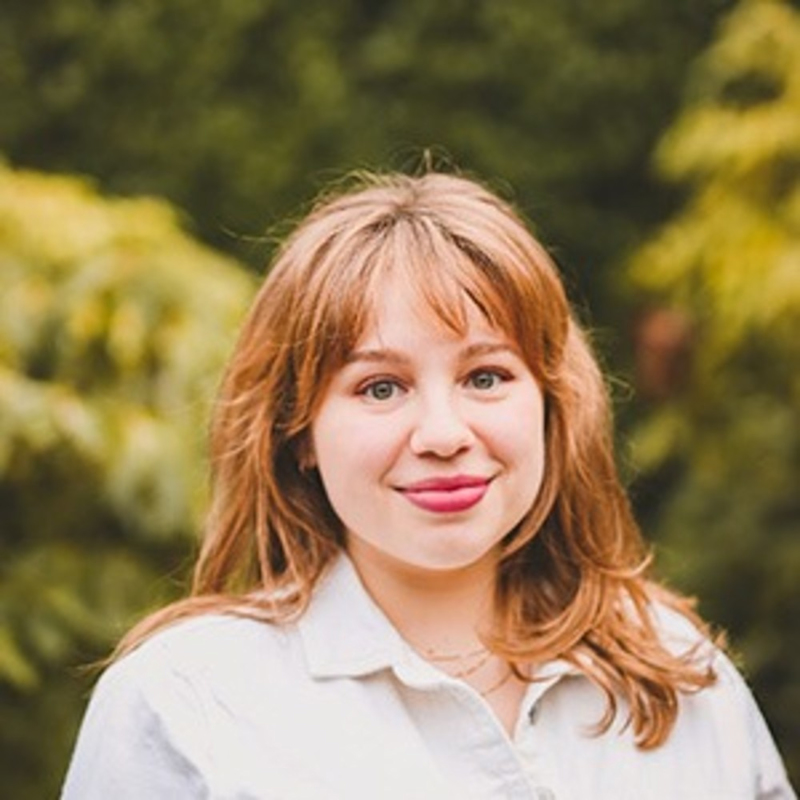 A person with shoulder-length hair smiles softly while wearing a light-colored collared shirt. The background is blurred greenery, providing a natural setting.