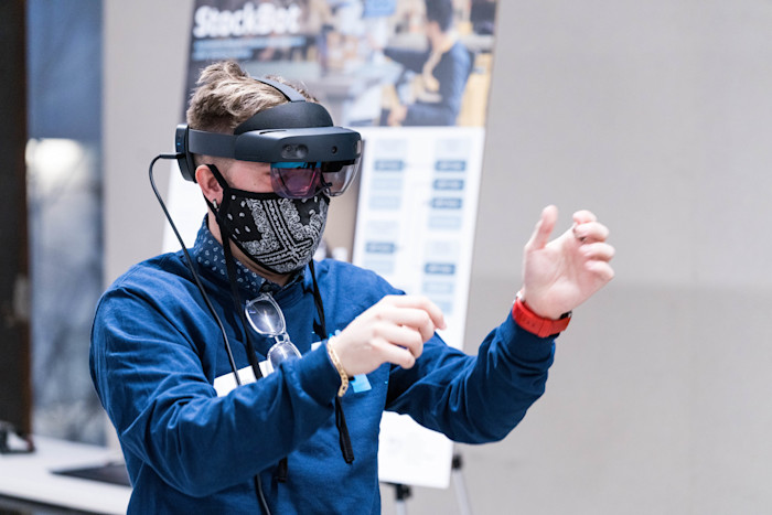 A person wearing a virtual reality headset and a patterned face mask gestures with both hands. They are dressed in a blue long-sleeve shirt. In the background, there is a blurred sign with text and images.