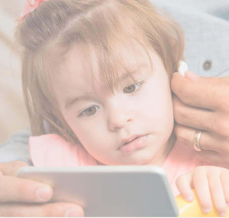 A toddler with brown hair watches a smartphone screen intently while an adult helps adjust a white earbud in her ear. The adults hand is visible, wearing a ring. The toddler wears a pink outfit and has a pink bow in her hair.