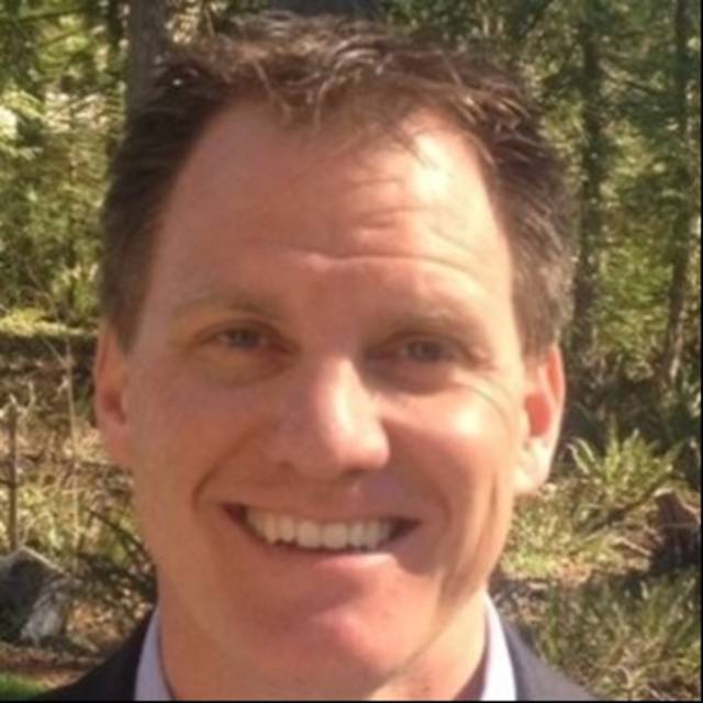 A man with short brown hair smiling outdoors. Trees and greenery are visible in the background.