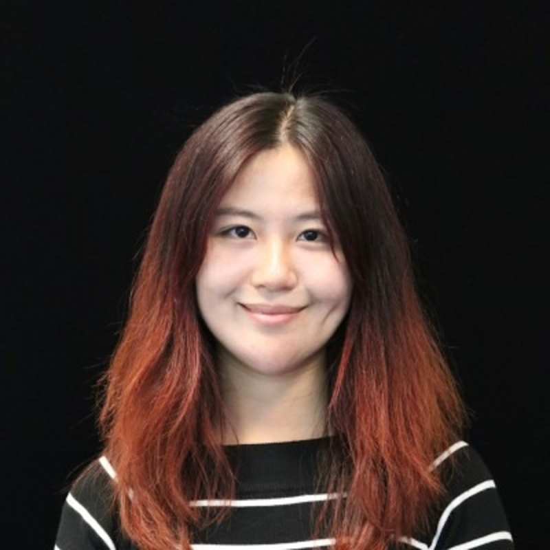 A person with shoulder-length hair, wearing a black striped shirt, smiles against a dark background.