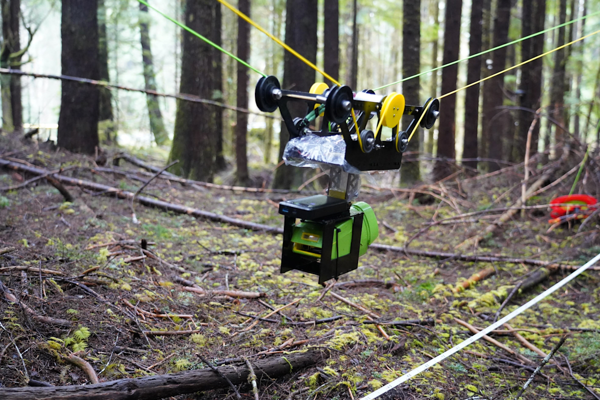A green and black robotic device is suspended by wires in a dense forest, equipped with wheels and pulleys. The ground is covered in moss and fallen branches, and the background is filled with tall trees.