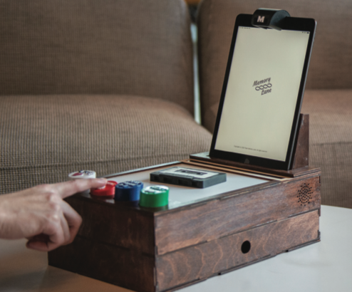 A person interacts with a device featuring colorful buttons and a tablet labeled Memory Good Zone. The device has a rustic wood finish and a cassette tape placed on it, set on a table with a couch in the background.