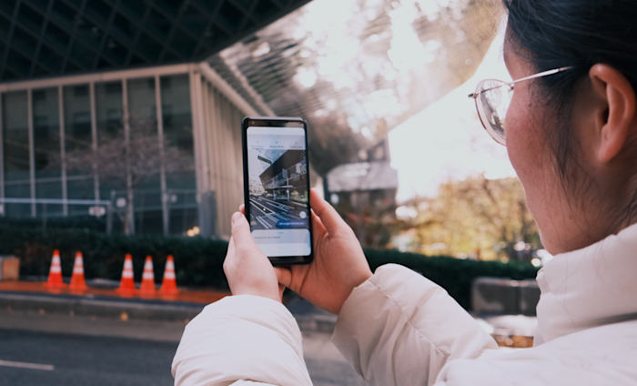 A person wearing a white jacket and glasses holds up a smartphone, using a map app while facing a modern glass building. Orange traffic cones are visible on the sidewalk. The scene suggests navigation or directions, with a focus on technology.