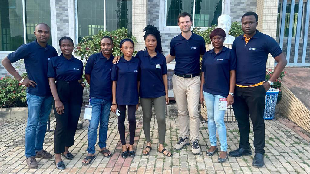 A group of eight people, wearing matching navy blue shirts, stand in a line outside a building. They are smiling and have name tags. The background features a brick walkway and plants.