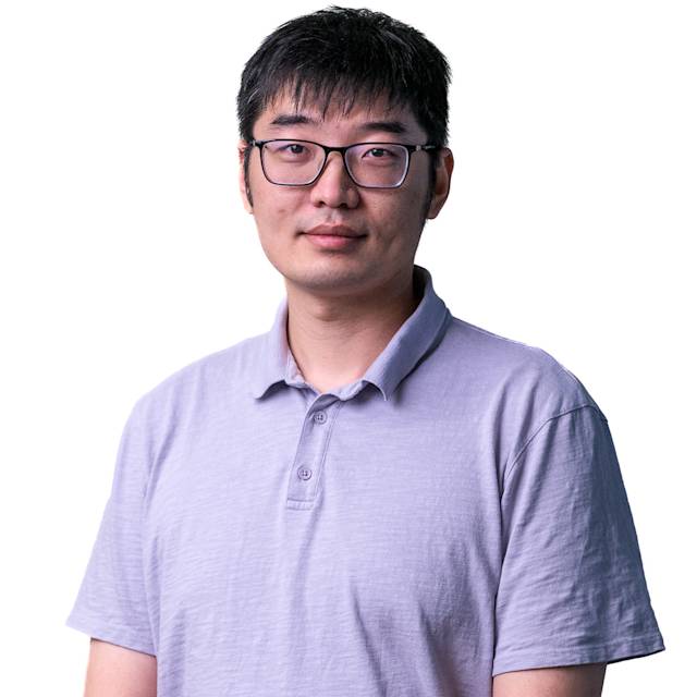 A person wearing glasses and a gray collared shirt stands in front of a light-colored, textured background. They look directly at the camera with a neutral expression.
