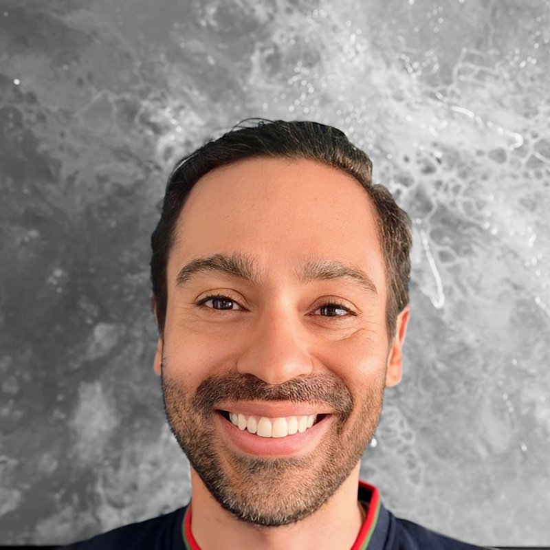 Smiling man with short dark hair and a beard stands in front of a textured gray background, appearing relaxed and cheerful.
