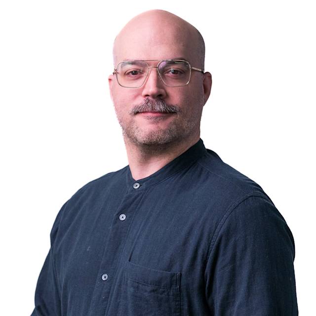 A person with a mustache, bald head, and glasses is wearing a gray button-up shirt. They are standing in front of a background with a pattern of small rectangles. The person is facing the camera with a slight smile.