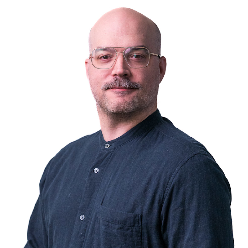 A person with a mustache, bald head, and glasses is wearing a gray button-up shirt. They are standing in front of a background with a pattern of small rectangles. The person is facing the camera with a slight smile.