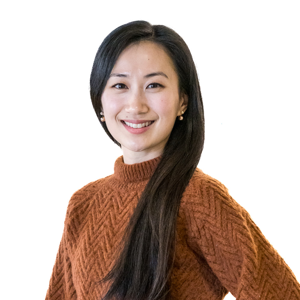 A person with long dark hair wearing a brown sweater stands smiling in front of a perforated metal wall. They have a confident posture with one hand on their hip.