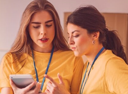 Two individuals wearing yellow shirts and blue lanyards are looking at a tablet. One gestures towards the screen while the other attentively observes. The background is softly blurred, suggesting an indoor setting.