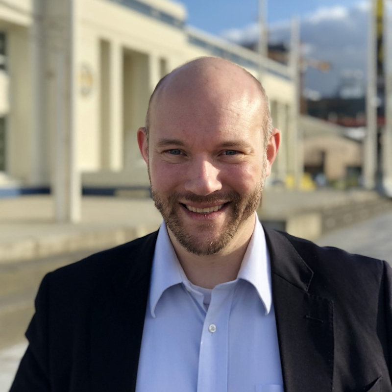 A person with a shaved head and beard is smiling at the camera. They are wearing a white shirt and a dark suit jacket. The background shows a building with columns under a clear sky.