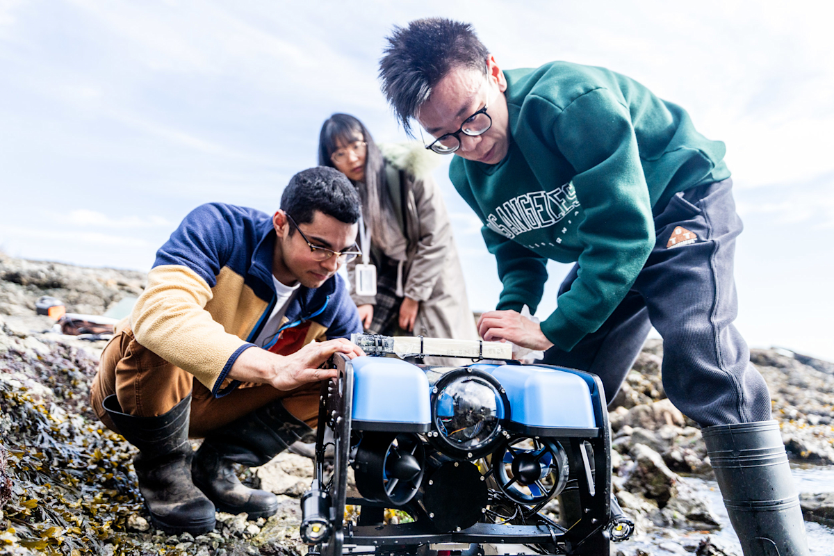 Three people on a rocky shore examine a small blue underwater drone. Two kneel beside it, focusing intently, while the third person stands in the background. They are surrounded by seaweed under a cloudy sky.