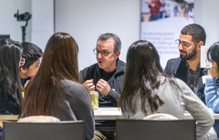A group of people sit around a table discussing. A person in the center is speaking, and others are listening attentively. A camera is set up in the background, capturing the meeting.