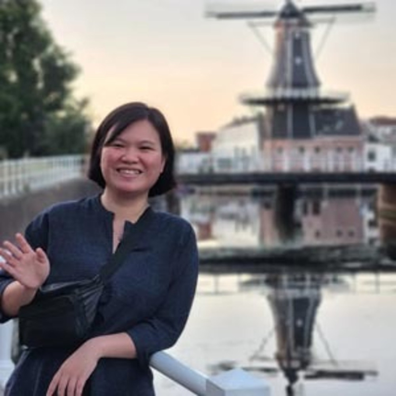 A person with short hair stands by a canal, smiling and waving. They wear a dark outfit and a crossbody bag. In the background, there is a windmill and a railing along the water. The sky is clear.