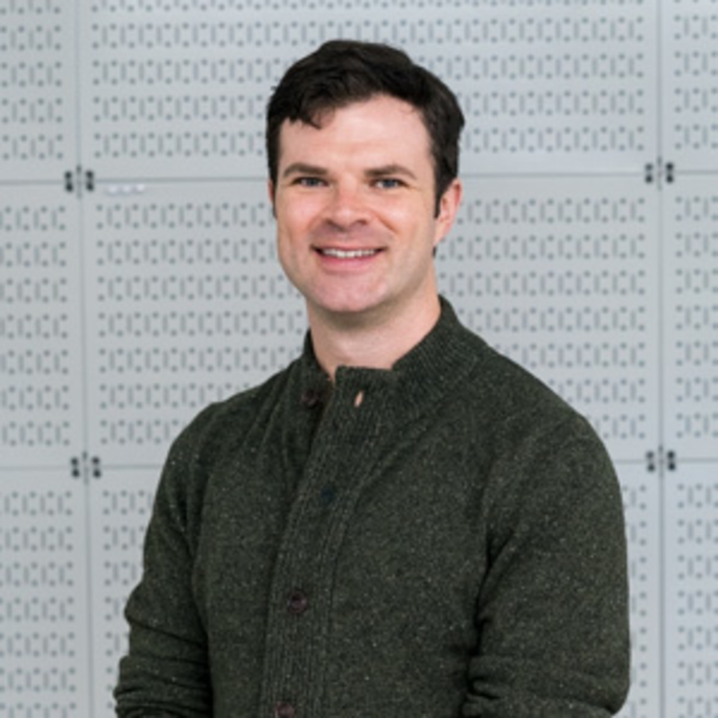 A person with short dark hair smiles at the camera, wearing a dark green sweater. The background features a light gray patterned wall.