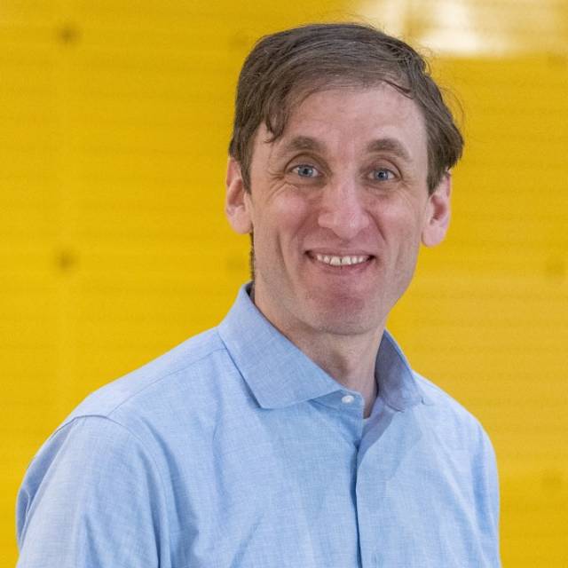 Man smiling, wearing a light blue button-up shirt, stands in front of a bright yellow background.