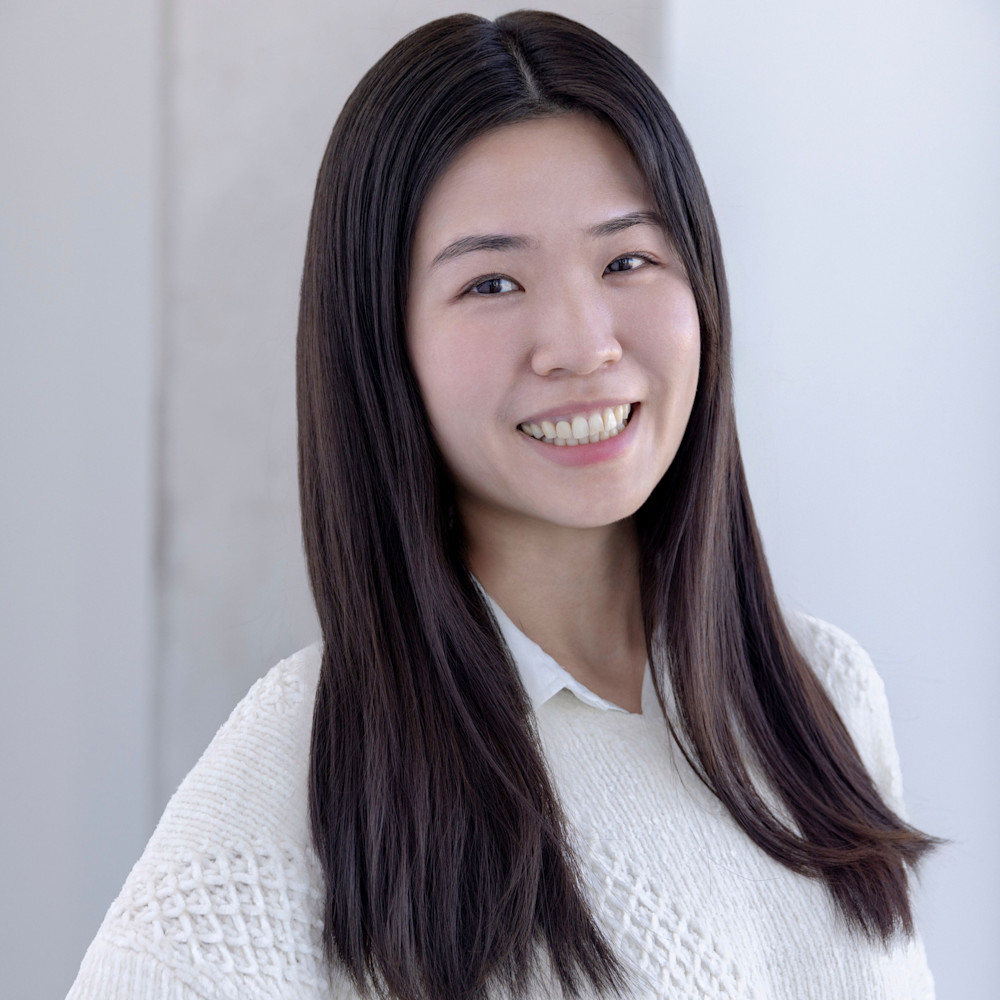 A person with long, straight hair smiles at the camera. They are wearing a light-colored, textured sweater over a collared shirt. The background is softly blurred, featuring a white wall.