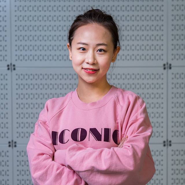 A person with dark hair is smiling and standing with crossed arms, wearing a pink sweatshirt with the word ICONIC on the front. The background is a wall with a pattern of rectangular shapes.