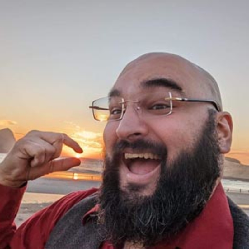 A bearded person with glasses is smiling widely on a beach at sunset. They point towards the sunset, which features a clear sky and a distant rock formation. They are wearing a red shirt and dark vest.