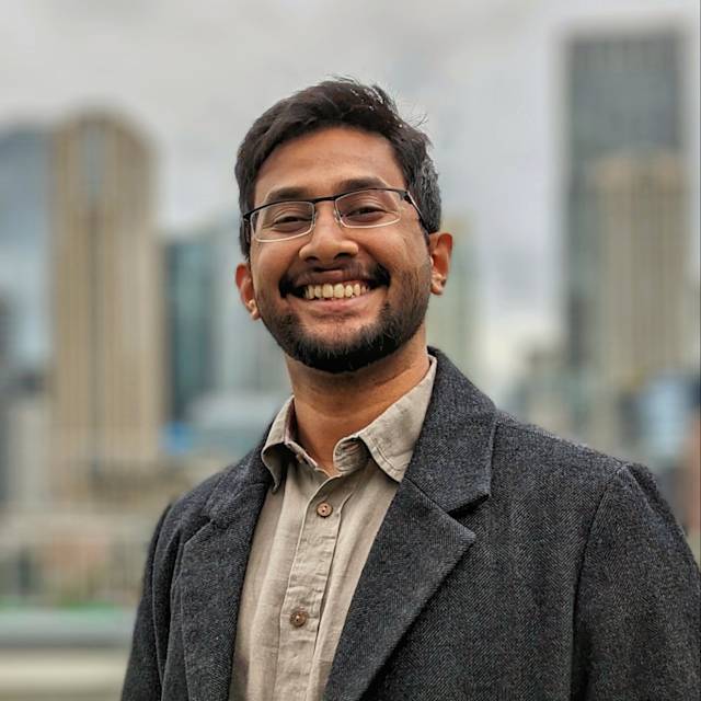 Smiling man wearing glasses, a gray coat, and a button-up shirt stands in front of a blurred urban skyline with tall buildings.