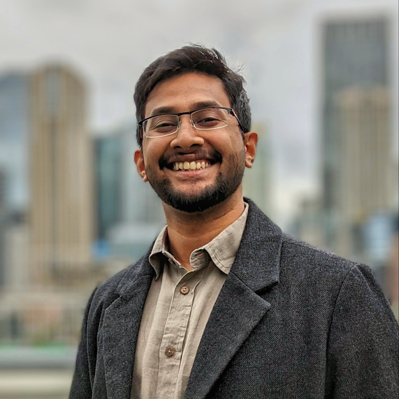 Smiling man wearing glasses, a gray coat, and a button-up shirt stands in front of a blurred urban skyline with tall buildings.