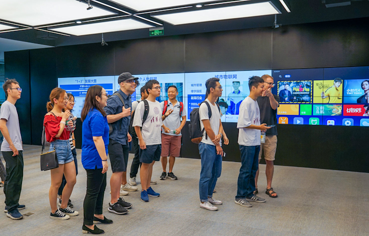 A group of people stands in front of a large digital screen displaying various apps and media content. They appear engaged and are casually dressed. The setting is modern with sleek, dark walls and bright lighting overhead.