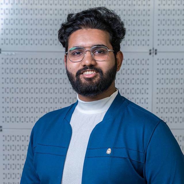 A person with dark hair, a beard, and glasses is smiling while wearing a blue jacket over a white shirt. The background consists of a patterned white wall.