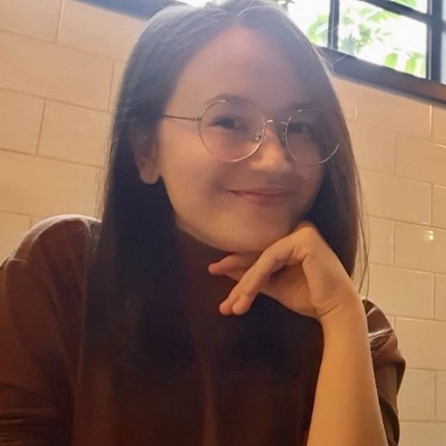 A person with long, dark hair and glasses is smiling, resting their chin on their hand. They are sitting indoors against a tiled wall with a window above. The lighting is warm and soft.
