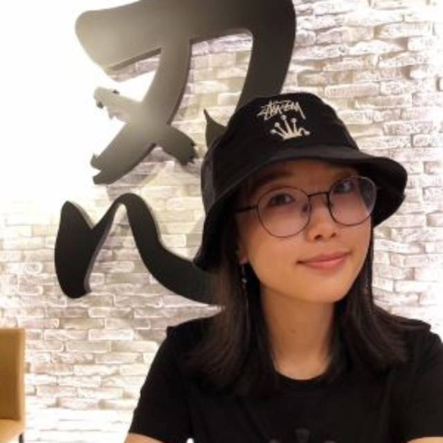 A person wearing glasses, a black bucket hat, and a black shirt sits at a table. They are smiling slightly. Behind them is a brick wall with large black stylized text or artwork.