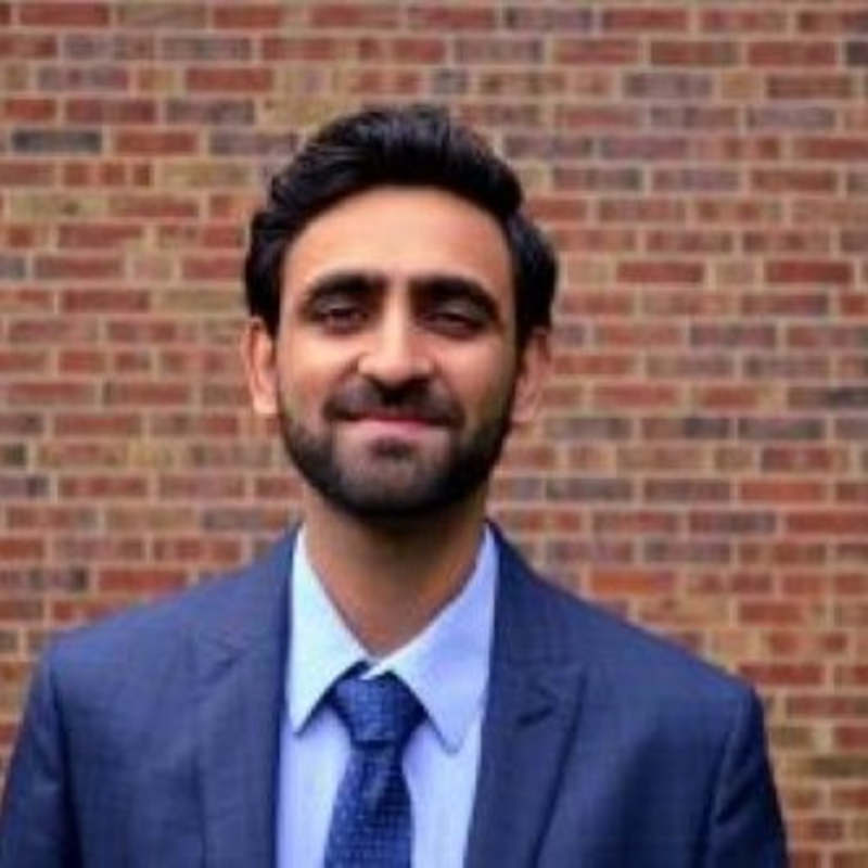 A man with dark hair and a beard is wearing a blue suit and tie, standing in front of a brick wall. He is smiling slightly at the camera.