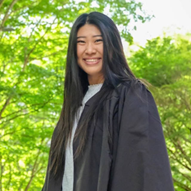 A person with long dark hair is smiling while wearing a graduation gown. The background features bright green trees and sunlight filtering through the leaves.