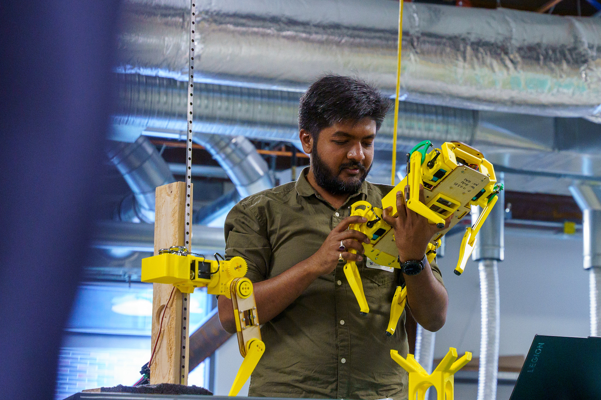 A person with a beard and short hair works on a yellow robotic dog in an industrial setting. Ventilation ducts are visible in the background. The person is wearing a green shirt and is focused on assembling or inspecting the robot.