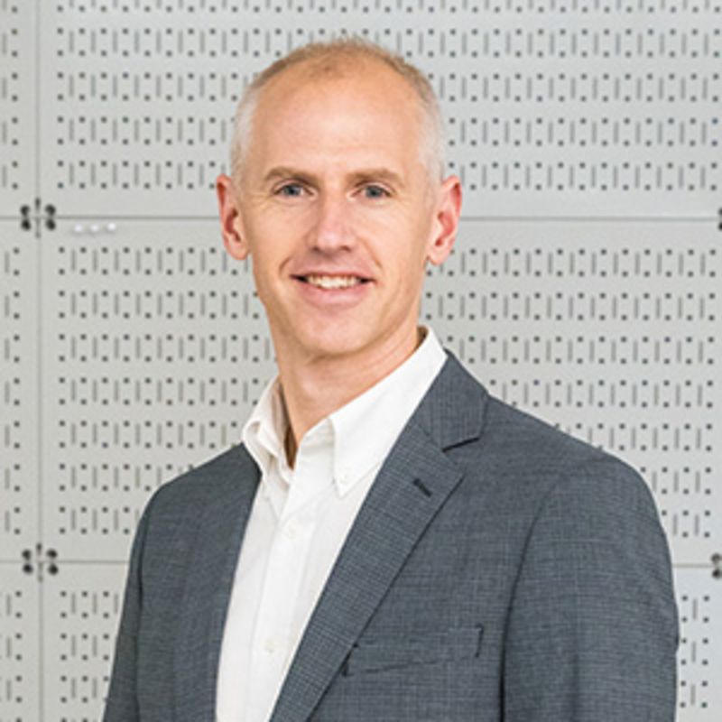 A person with short, light hair is smiling at the camera, dressed in a dark gray blazer over a white shirt. The background is a perforated white wall with a grid pattern.