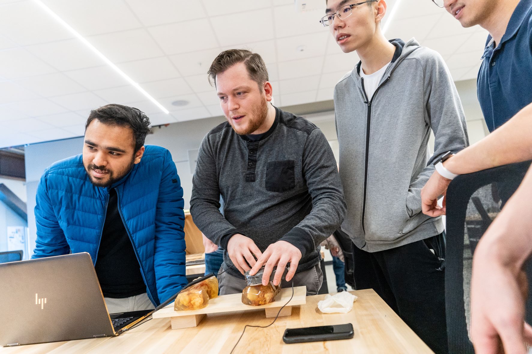 A group of four people, three men and one person partially visible, gather around a laptop on a wooden table with sandwiches. One man is adjusting the sandwiches while others watch. They seem focused on the task.
