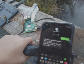 A person holds a smartphone displaying a text message. Nearby, theres an electronic circuit board connected to a laptop. The setup is on a wooden surface outdoors, with a blurred background of grass and water.